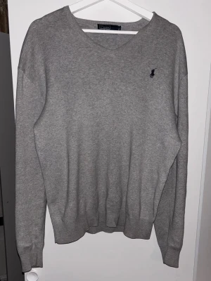 Grå v-ringad tröja från Polo Ralph Lauren - Klassisk grå v-ringad tröja från Polo Ralph Lauren med diskret broderad logga på bröstet. Tröjan har långa ärmar och ribbade muddar vid ärmslut och nederkant. Perfekt för en stilren och enkel look. Upphämtning tillgänglig i Stockholm