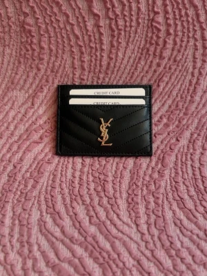 Svart kortfodral från Saint Laurent - Snyggt svart kortfodral från Saint Laurent i quiltat läder med guldfärgad YSL-logga framtill. Fodralet har flera kortfack och en stilren, minimalistisk design som passar perfekt i fickan. Perfekt accessoar för dig som gillar lyxiga detaljer.