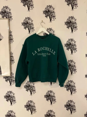 Grön sweatshirt från Gina Tricot - Mörkgrön sweatshirt från La Rochelle med trycket 'Vélo Sport Club' i vitt framtill. Tröjan har rund halsringning, ribbade muddar vid ärmslut och nederkant samt en avslappnad passform. Perfekt för en sportig och chill stil.