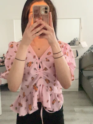 Rosa blommig blus med snörning - Supersöt rosa blus med blommigt mönster och volangkant. Blusen har v-ringning, puffiga korta ärmar och snörning framtill som ger en snygg passform. Perfekt för dig som gillar romantisk och trendig stil.