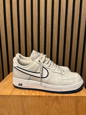 Nike Air Force 1 vita sneakers - Fräscha vita Nike Air Force 1 sneakers med marinblå detaljer och klassisk swoosh på sidan. Skorna har perforerad tå, platt sula med AIR-dämpning.