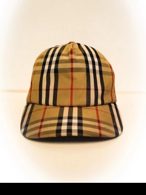 Berberry Keps - Snygg keps från Burberry i beige med det ikoniska rutmönstret i svart, vitt och rött. Kepsen har böjd skärm och justerbar rem bak. Tillverkad i bomull för en bekväm och stilren look.