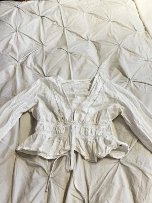 Vit blus med spetsdetaljer och knytband - Snygg vit blus med långa ärmar, v-ringning och dekorativa spetsdetaljer längs bröstet och ärmarna. Blusen har resår i midjan och ett knytband framtill för en feminin siluett. Tillverkad i ett lätt och luftigt tyg, perfekt för en trendig look.