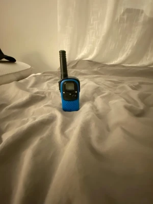 Blå walkie talkie med svart antenn - En smidig walkie talkie i blå plast med svart antenn och svart frontpanel. Den har en liten display och knappar på framsidan för enkel användning. Perfekt för kommunikation på kortare avstånd, till exempel på äventyr eller festival.