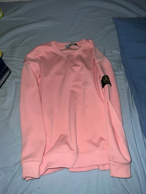 Stone Island - Säljer en ljusrosa sweatshirt från Stone Island med klassisk rund hals och ribbade muddar. Tröjan har den ikoniska svarta logotypen på vänster ärm och är gjord i mjukt bomullsmaterial. Perfekt för en avslappnad och trendig look.