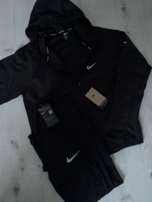 Nike "miller" set - Snyggt svart träningsset från Nike med jacka och byxor. Jackan har huva, dragkedja och reflekterande detaljer samt Nike-logga på bröstet. Byxorna har resår i midjan och Nike-logga på benet. Perfekt för träning utomhus.