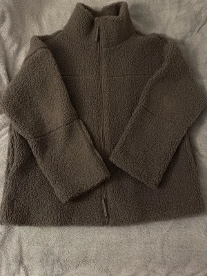 Brun teddyjacka med dragkedja - Mysig brun teddyjacka med hög krage och hel dragkedja framtill. Jackan har en boxig passform och är gjord i fluffigt teddy-material som håller dig varm. Perfekt för kalla dagar och enkel att matcha med olika outfits.