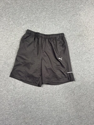 Svarta träningsshorts från Puma - Snygga svarta träningsshorts från Puma med elastisk midja och snörning. Shortsen har en diskret logga på ena benet och reflekterande detaljer. Tillverkade i lätt och snabbtorkande syntetmaterial, perfekta för aktiva dagar.