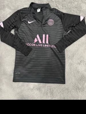 Svart PSG träningströja från Nike - Snygg svart långärmad träningströja från PSG med rosa detaljer och logga. Tröjan har halv dragkedja, diskret mönster och trycket 'Accor Live Limitless' på bröstet. Perfekt för fotbollsträning eller chill. Tillverkad i snabbtorkande material.