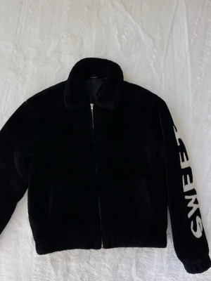 Svart teddyjacka med SWEET-tryck - Mysig svart teddyjacka med dragkedja framtill och två fickor. Jackan har en klassisk krage och ett stort vitt SWEET-tryck längs ena ärmen