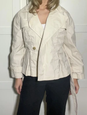 Beige trenchcoat med bälte - Stilren beige trenchcoat med bred krage och knytband i midjan. Jackan har en knappdetalj framtill och markerad midja för en snygg siluett. Tillverkad i ett lätt tyg som passar perfekt till vår och höst. Klassisk design från & other stories.
