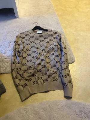 Beige tröja med GG-mönster från Gucci - Snygg beige tröja från Gucci med klassiskt GG-monogram över hela plagget. Rund halsringning och ribbade muddar vid ärmslut och nederkant. Perfekt för dig som gillar exklusiv streetstyle och vill sticka ut.