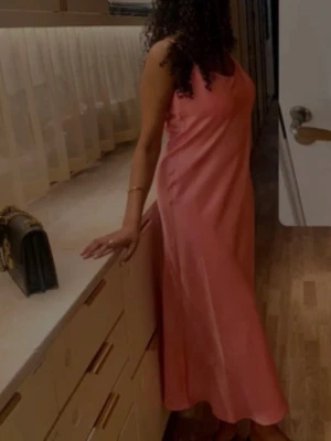 Rosa långklänning från Kappahl - Säljer en elegant rosa långklänning från Kappahl i glansigt satinliknande material. Klänningen har smala axelband och en rak, enkel siluett som ger en stilren look. Perfekt för dig som vill sticka ut med en färgstark och trendig outfit.