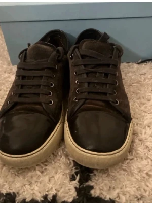 Svarta sneakers från Lanvin - Snygga svarta sneakers från Lanvin med ovandel i mocka och tåparti i blankt läder. Skorna har svarta skosnören, metallöljetter och en kontrasterande vit sula. Perfekta för en stilren och trendig look.