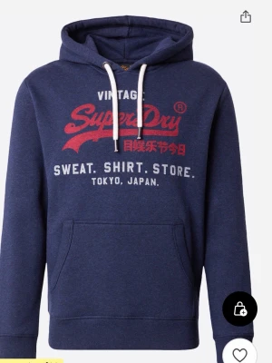 Blå Superdry hoodie med tryck - Mörkblå hoodie från Superdry med rött och vitt tryck på bröstet. Klassisk huva med vita snören och stor känguruficka framtill. Mjuk bomullsblandning och ribbade muddar vid ärmslut och nederkant. Perfekt för en avslappnad streetstyle.
