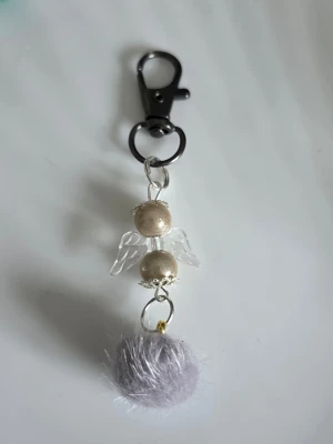 Nyckelring med ängel och fluffboll reflex pärlor - Söt nyckelring med en ängel gjord av pärlor och genomskinliga vingar samt en mjuk, ljusgrå fluffboll. Metallhake i svart ger en cool kontrast. Perfekt att hänga på väskan eller nycklarna för en personlig touch.