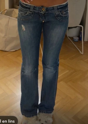 Blå bootcut jeans med slitningar - Säljer ett par blå bootcut jeans med låg midja och slitna detaljer på låren och knäna. Jeansen har klassiska fem fickor och kontrastsömmar. Perfekta för en avslappnad och trendig look.