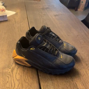 Nocta hot step air terra - Unika svarta sneakers från Nike med coola gula detaljer och en chunky sula i gult. Skorna har snörning, rund tå och är tillverkade i syntetmaterial med skinnliknande yta. Perfekt för dig som vill sticka ut med en sportig och modern look.