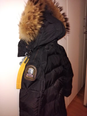 Svart Parajumpers dunjacka Longbear med päls - Svart lång dunjacka Longbear från Parajumpers med fluffig brun päls runt huvan och ikonisk gul rem på axeln. Jackan har patch på ärmen, dragkedja framtill och stora fickor. Perfekt för kalla dagar och riktigt snygg streetstil.