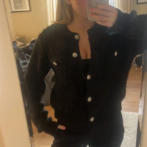 Svart kofta med silvriga knappar - Mysig svart kofta från H&M i storlek S. Koftan är stickad med fluffig textur och har silvriga dekorativa knappar framtill samt på de två bröstfickorna. Rund halsringning och ribbade muddar vid ärmslut och nederkant. Perfekt för lager-på-lager under kyliga dagar.