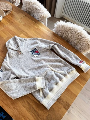 Grå New York Rangers Cardigan - Säljer en grå stickad cardigan med New York Rangers-logga på bröstet och tryck på ärmen. Tröjan har ribbade muddar, ficka framtill och två knappar vid halsen. Perfekt för dig som gillar hockey och vill ha en sportig look.