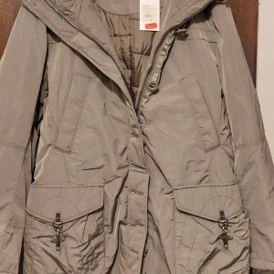 Beige parkajacka med huva - Snygg beige parkajacka med huva och stora fickor framtill. Jackan har dragkedja och knappstängning, samt coola detaljer med spännen på fickorna. Perfekt för dig som gillar en clean och praktisk stil.