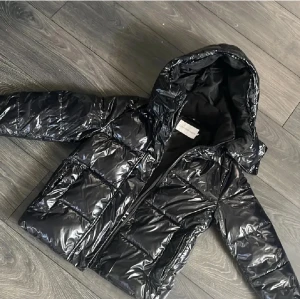 Svart pufferjacka Calvin Klein Jeans - Snygg svart pufferjacka från Calvin Klein Jeans med glansig finish och stor huva. Jackan har CK-logga på bröstet, dragkedja framtill och quiltad design. Perfekt för kalla dagar och har en modern, urban vibe.