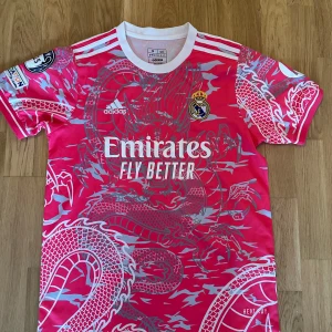 Real Madrid rosa Bellingham tröja M - Säljer en fet Real Madrid fotbollströja från Adidas i rosa med grått drakmönster, Bellingham #5 på ryggen och vita detaljer. Tröjan har korta ärmar, rund hals och broderat klubbmärke. Materialet är lätt och andas, perfekt för match eller träning.