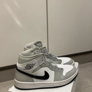 Nike Air Jordan 1 Mid Grey Fog - Nike Air Jordan 1 Mid i färgen Grey Fog med svart swoosh och vita paneler. Klassisk high-top siluett med snörning och perforerad tå. Skorna har en stilren och clean look med gråa och vita detaljer, samt ikoniska Air Jordan-loggan på sidan.