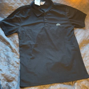 Svart pikétröja från Lacoste M - Svart pikétröja från Lacoste i klassisk regular fit. Tröjan har korta ärmar, krage med tre knappar och den ikoniska broderade krokodilen på bröstet. Materialet är mjuk bomull som andas och känns skönt mot huden. Perfekt för dig som gillar stilren och enkel design.