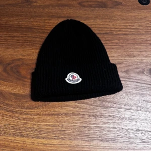 Svart ribbstickad mössa från Moncler - Svart ribbstickad mössa från Moncler med klassisk uppvikt kant och broderad logotyp framtill. Tillverkad i mjukt ullmaterial som håller dig varm och snygg. Perfekt accessoar för kyliga dagar och en clean look.