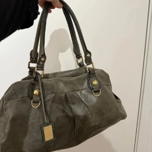 Grön handväska från Dolce & Gabbana - Snygg grå handväska från Dolce & Gabbana i skinn med guldfärgade metalldetaljer och dubbla handtag. Väskan har en rymlig insida med dragkedja och dekorativa veck framtill. Perfekt för dig som vill ha en lyxig och stilren accessoar.