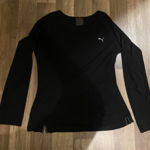 Svart långärmad träningströja Puma - Svart långärmad träningströja från Puma med diskret logga på bröstet. Tröjan har rund hals och är tillverkad i mjukt, stretchigt syntetmaterial som andas. Små slitsar med dragkedja i nederkant ger extra stil och rörelsefrihet.