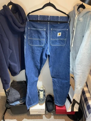 Blå Carhartt jeans med raka ben - Säljer ett par klassiska blå jeans från Carhartt med raka ben och kontrasterande vita sömmar. Byxorna har stora bakfickor, varav en med Carhartt-logga, och praktiska detaljer som förstärkta sömmar. Perfekt för dig som gillar streetwear och funktionell stil.
