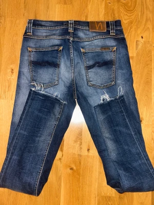 Mörkblå jeans från Nudie med slitningar - Snygga mörkblå jeans från Nudie med klassiska femficksdetaljer och orangea sömmar. Jeansen har slitna partier och råa kanter vid bensluten för en cool, avslappnad look. Passformen är rak och materialet är robust denim.
