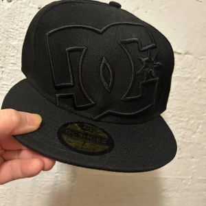 Svart DC Shoes keps 7 (55,8 cm) - Snygg svart keps från DC Shoes med stort broderat DC-logo framtill. Klassisk flat brim och cool streetstil. Kepsen är i storlek 7 (55,8 cm) och har detaljer i svart ton-i-ton. Perfekt för dig som gillar skate eller streetwear.
