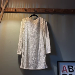 Vit paljettklänning med svart rosett - Säljer en glittrig vit kortklänning från JDY, täckt med paljetter och med långärmad design. Klänningen har en snygg svart rosett vid halsen som ger en extra cool touch. Perfekt för dig som vill sticka ut med stil och glitter. 