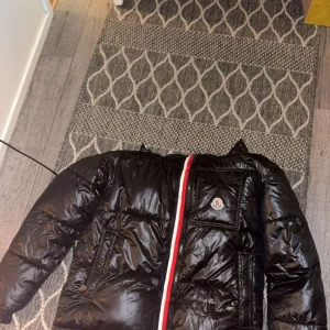 Svart Moncler pufferjacka med huva - Svart pufferjacka från Moncler med glansig finish, huva och ikonisk logga på bröstet. Jackan har en rödvit rand längs dragkedjan och flera fickor. Insidan har en cool serietidningspatch. Perfekt för dig som vill ha en snygg och varm vinterjacka.