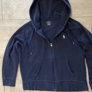 Marinblå hoodie från Polo Ralph Lauren - Snygg marinblå hoodie från Polo Ralph Lauren med dragkedja och klassisk logga broderad på bröstet. Tröjan har huva med snörning, två fickor fram och ribbade muddar vid ärmslut och nederkant. Perfekt för chill dagar och streetstyle.
