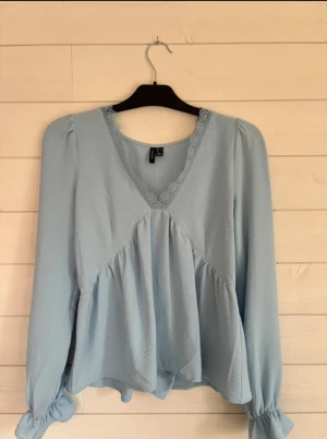 Ljusblå blus med spetsdetalj - Supersöt ljusblå blus med v-ringning och spetsdetalj runt halsen. Blusen har puffiga långärmar med volang vid ärmslut och en luftig, peplumliknande form. Perfekt för dig som gillar romantiska och feminina plagg.