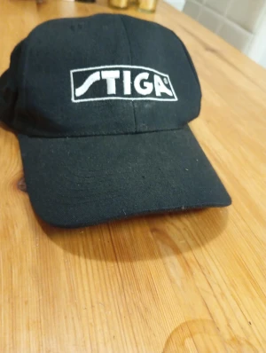 Svart keps från STIGA - Svart keps med klassisk böjd skärm och vit broderad STIGA-logga framtill. Tillverkad i bomull, perfekt för dig som gillar sportig stil och vill ha en enkel accessoar med tydlig branding.