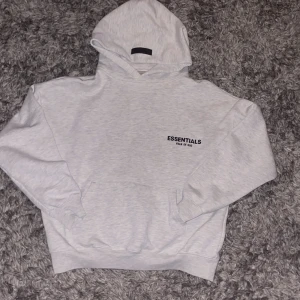 Ljusgrå Essentials Fear of God hoodie - Säljer en ljusgrå hoodie från Essentials Fear of God med svart logotyptryck både fram och bak. Tröjan har huva, känguruficka och ribbade muddar. Perfekt för dig som gillar streetwear och vill ha en clean look.
