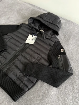 Moncler cardigan  - Snygg svart cardigan från Moncler med stickade ärmar och ribbade muddar. Jackan har huva, dragkedja framtill och en extra ficka med dragkedja på ärmen. Klassisk Moncler-logga på vänster ärm. Perfekt för kyliga dagar och riktigt stilren look.