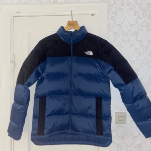 Blå dunjacka från The North Face M - Säljer en blå och svart dunjacka från The North Face i storlek M. Jackan har hög krage, dragkedja framtill och två svarta fickor. Klassisk pufferstil med quiltad design och lång ärm, perfekt för kalla dagar. den är använd en gång köptes 17 november från butik alltså inprincip helt ny