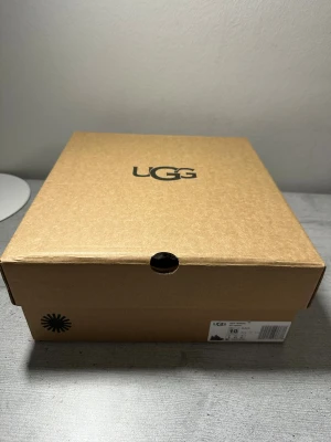 Svarta sneakers från UGG med foder - Säljer ett par svarta UGG sneakers med breda snören, grov sula och mysigt vitt foder på insidan. Ovandelen är i en mix av mocka och textil, vilket ger en schysst kontrast. Perfekta för dig som vill ha både stil och komfort under kyligare dagar. Skorna är aldrig använda☺️