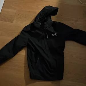 Svart vindjacka från Under Armour - Svart vindjacka från Under Armour med huva och dragkedja framtill. Jackan har en sportig look med logga på bröstet och praktiska fickor. Perfekt för blåsiga dagar och outdoor-aktiviteter.