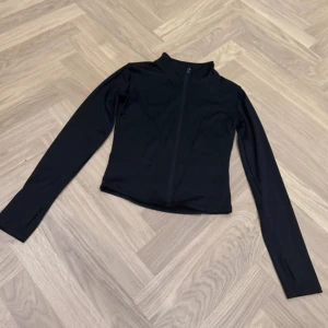 Svart sportig jacka från Stronger - Svart, figurnära sportjacka från Stronger med hel dragkedja framtill och hög krage. Jackan är tillverkad i stretchigt syntetmaterial och har långa ärmar med tumhål. Perfekt för träning eller som ett extra lager.