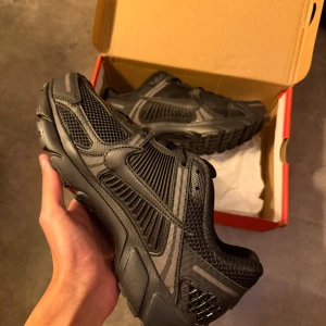 Nike Zoom Vomero 5 Svarta sneakers - Nike Zoom Vomero 5 sneakers i helsvart färg med mesh och syntetpaneler, chunky sula och snygga detaljer på hälen. Ikonisk Swoosh på sidan och grovt mönstrad yttersula för extra grepp. Perfekt för dig som gillar tech-vibe och sportig stil.
