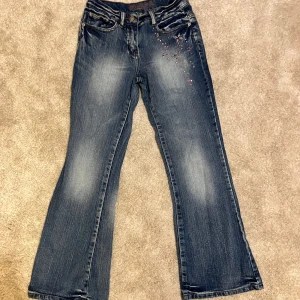 Bootcut jeans med rosa strass  - Snygga blå bootcut jeans med coola strassdetaljer i rosa och silver på framsidan och bakfickorna. Perfekta för dig som vill sticka ut lite extra. Använt men fint skick, enda defekten är att dragskedjan åker ner ganska lätt. Märket ser ut att vara ”onlyou” men kan inte hitta info om det förutom andra second hand kläder med samma märke. Justerbar midja