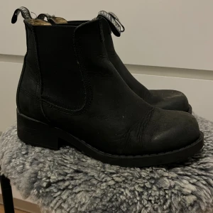 Svarta chelsea boots från Johnny Bulls - Snygga svarta chelsea boots från Johnny Bulls med grov sula och rund tå. Skorna är tillverkade i skinn och har elastiska paneler på sidorna för enkel på- och avtagning. Klassisk och stilren design med dragflik baktill och robust yttersula.
